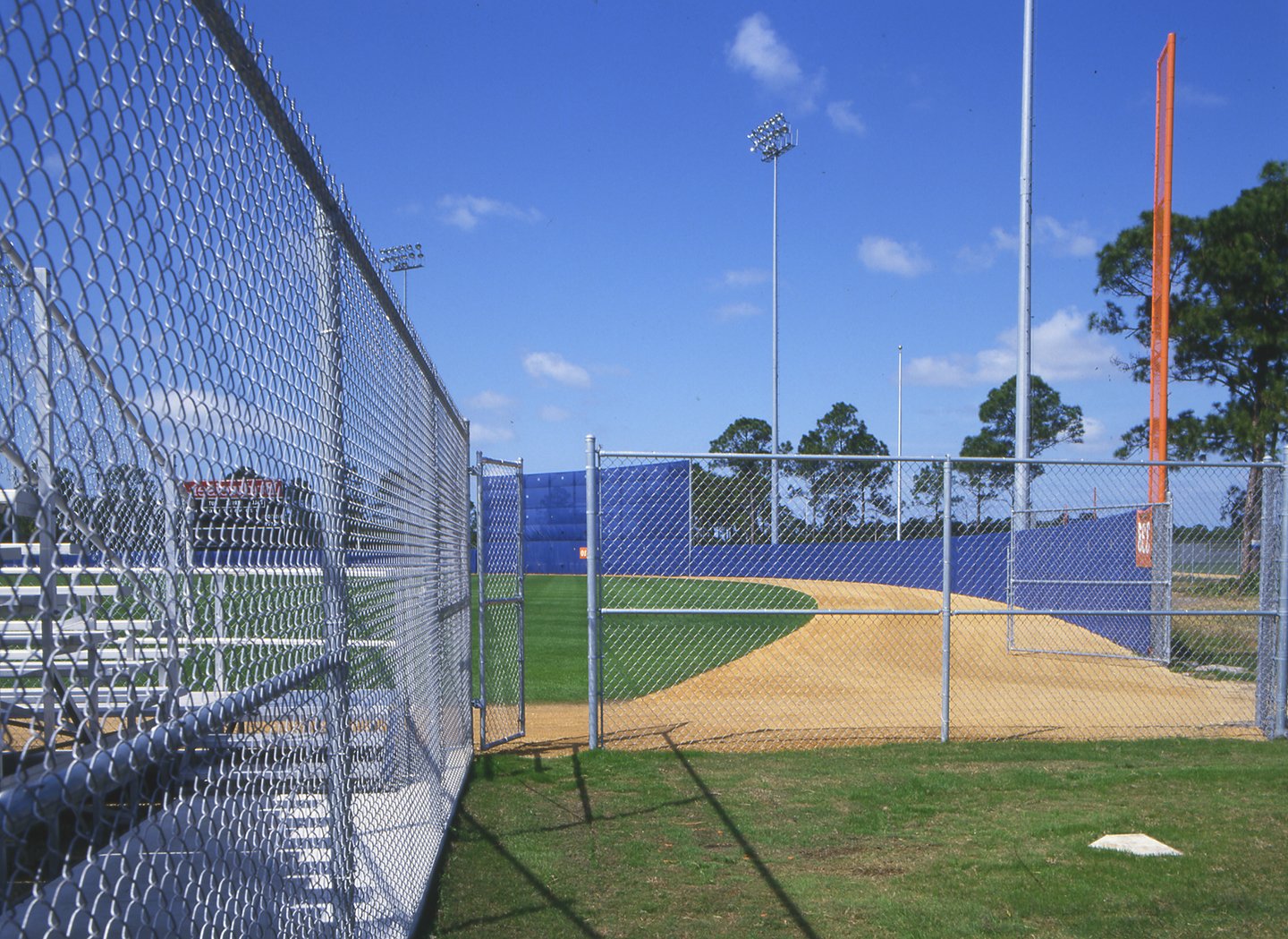 Galvanized chain link fence and gate surrounding a baseball field with light poles and blue outfield wall