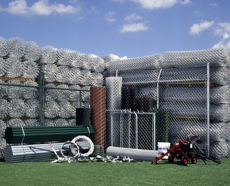Stacked rolls of chain link fabric, fence posts, rails, gates and fittings displayed together on a grass lawn