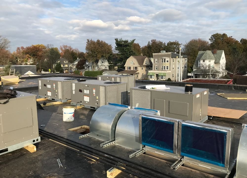 Rooftop HVAC units and metal ductwork with blue-covered openings installed on a flat commercial roof beside neighborhood houses