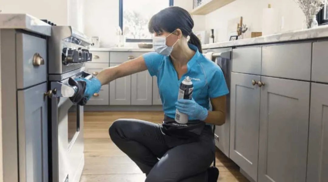 Masked cleaner in a teal uniform and blue gloves kneels in a kitchen and wipes down an oven
