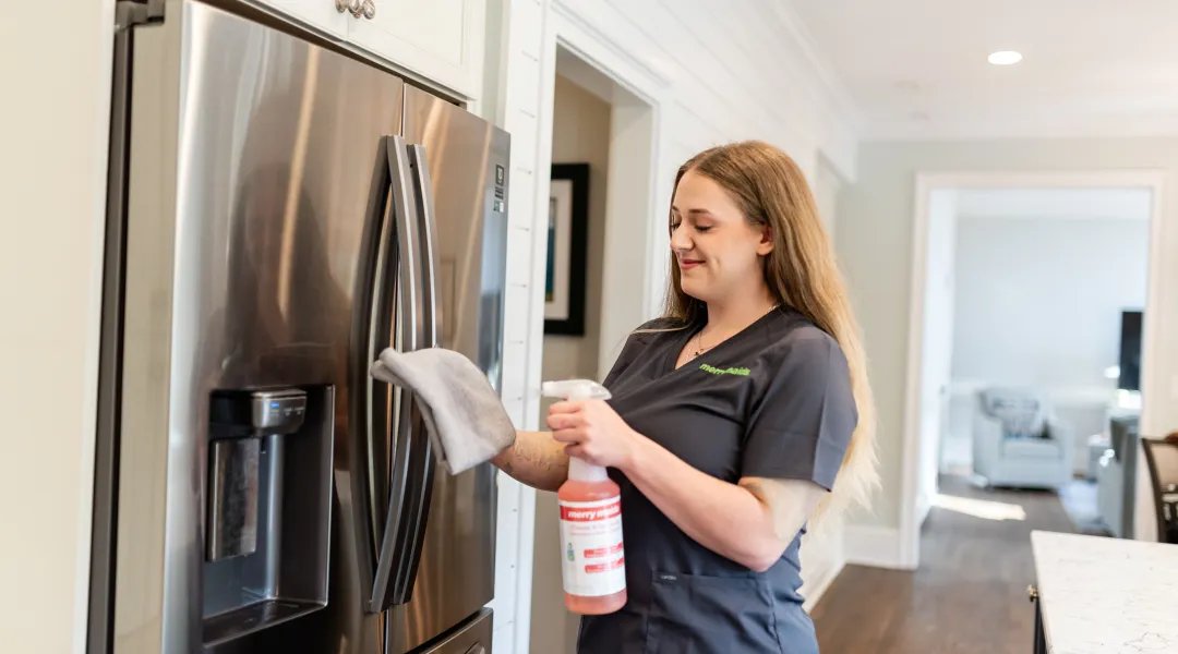Cleaner in a gray Merry Maids uniform wipes a stainless steel refrigerator door with a cloth and spray bottle