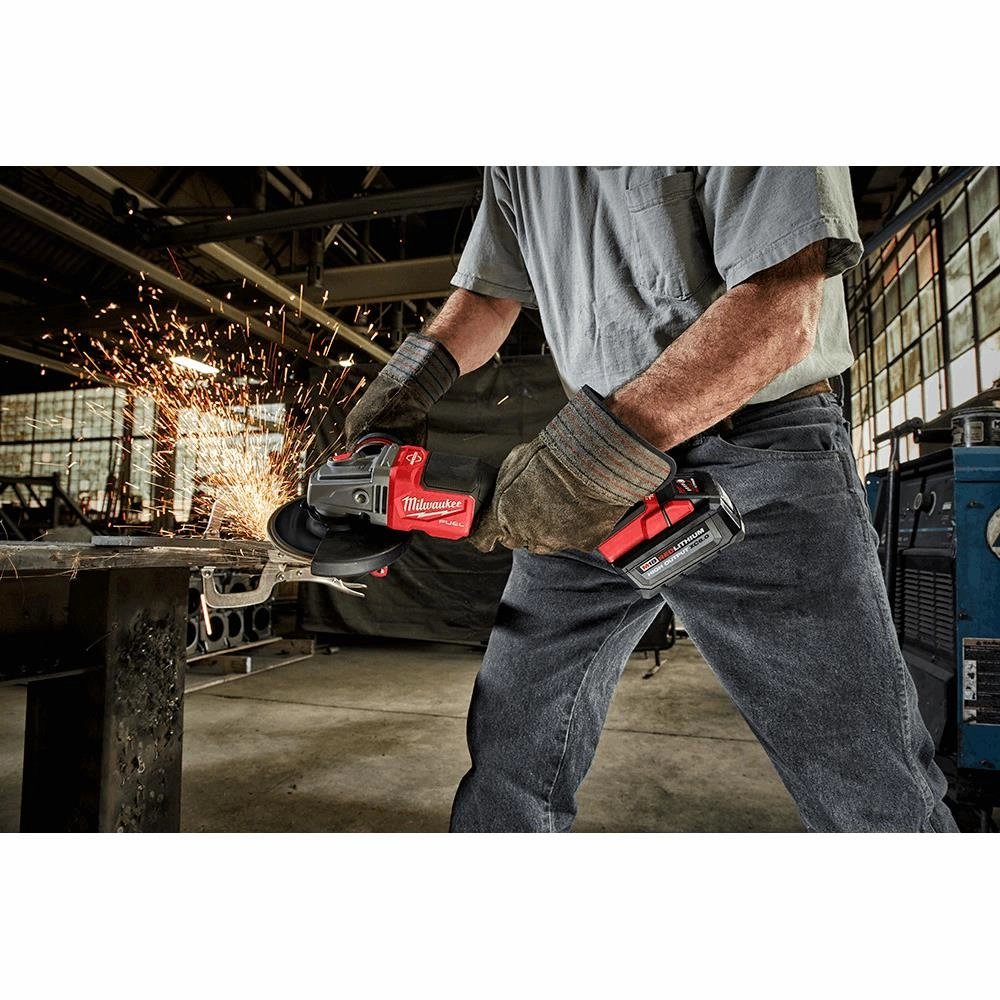 Worker in heavy gloves grinds a metal workpiece with a red Milwaukee cordless angle grinder, sparks flying in a shop