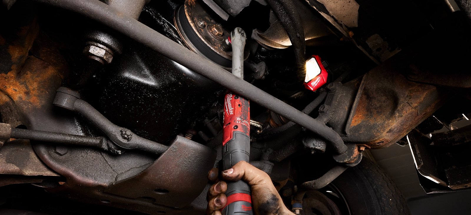 Grease-stained hand holding a red Milwaukee cordless ratchet beneath a vehicle, with a work light clipped to the undercarriage