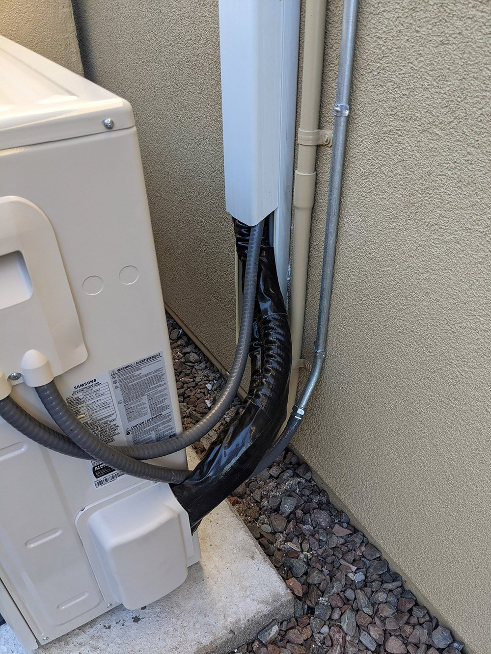Close-up of wrapped refrigerant lines and metal conduit running down a stucco wall from the back of an outdoor unit