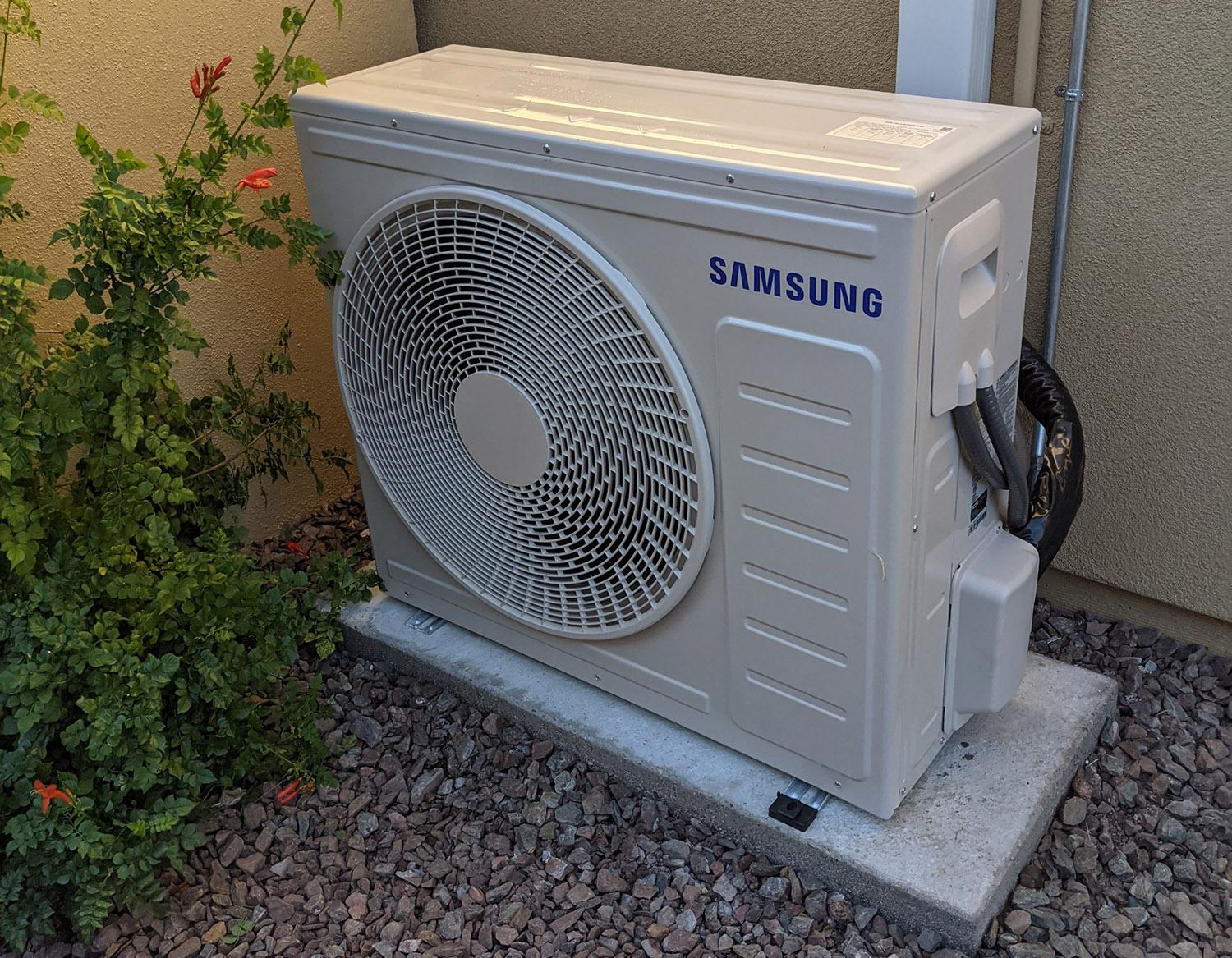 Samsung outdoor mini-split condenser on a concrete pad beside a stucco wall, with gravel and a flowering shrub nearby