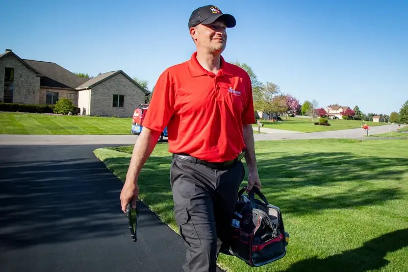 Technician in a red company polo and cap carries a tool bag up a suburban driveway, a van parked behind him