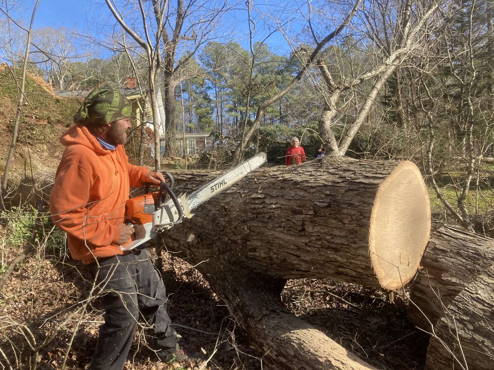 A man in an orange hooded sweatshirt and a green head wrap holds a Stihl chainsaw beside a large felled trunk in a wooded yard, as two people watch from behind the log