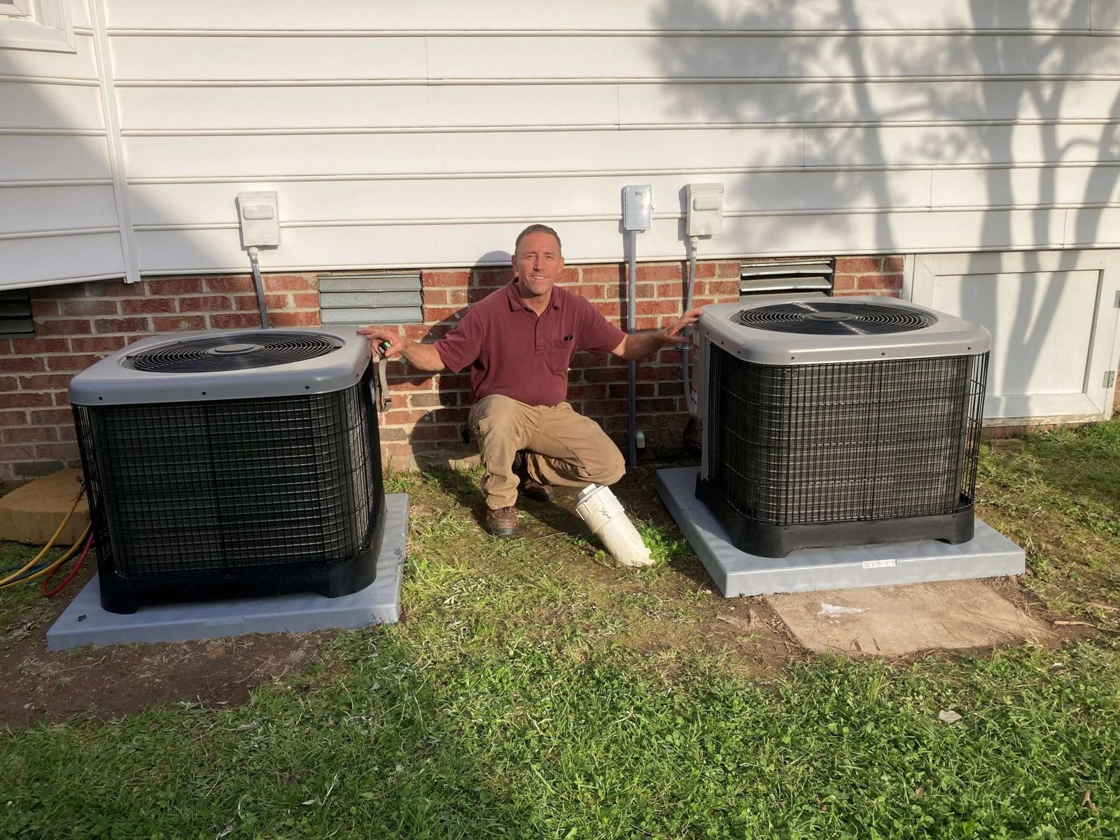 Technician in a maroon shirt crouches between two newly set outdoor condenser units on gray pads beside a brick home