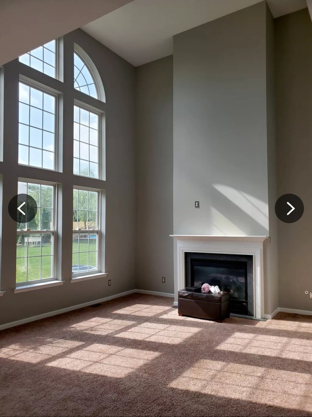 Freshly painted gray living room with a two-story fireplace wall, tall arched windows and sunlight across the carpet
