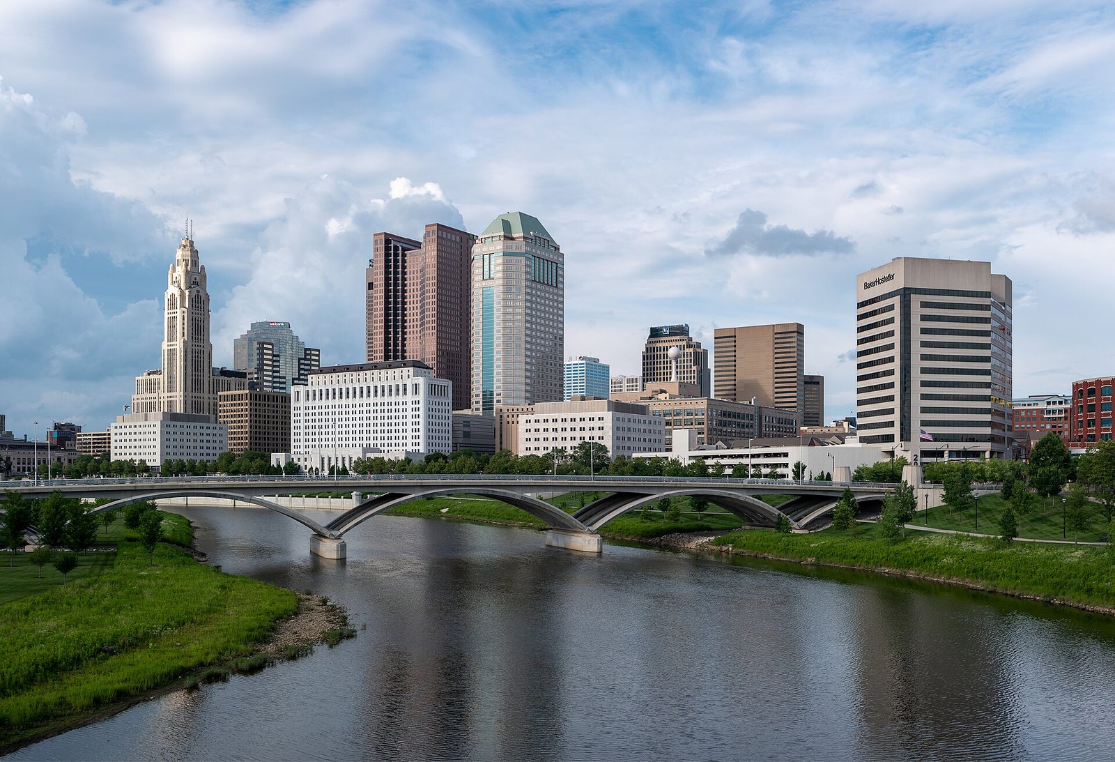 Columbus, Ohio skyline of office towers rising beside a river and a multi-arched bridge under a partly cloudy sky