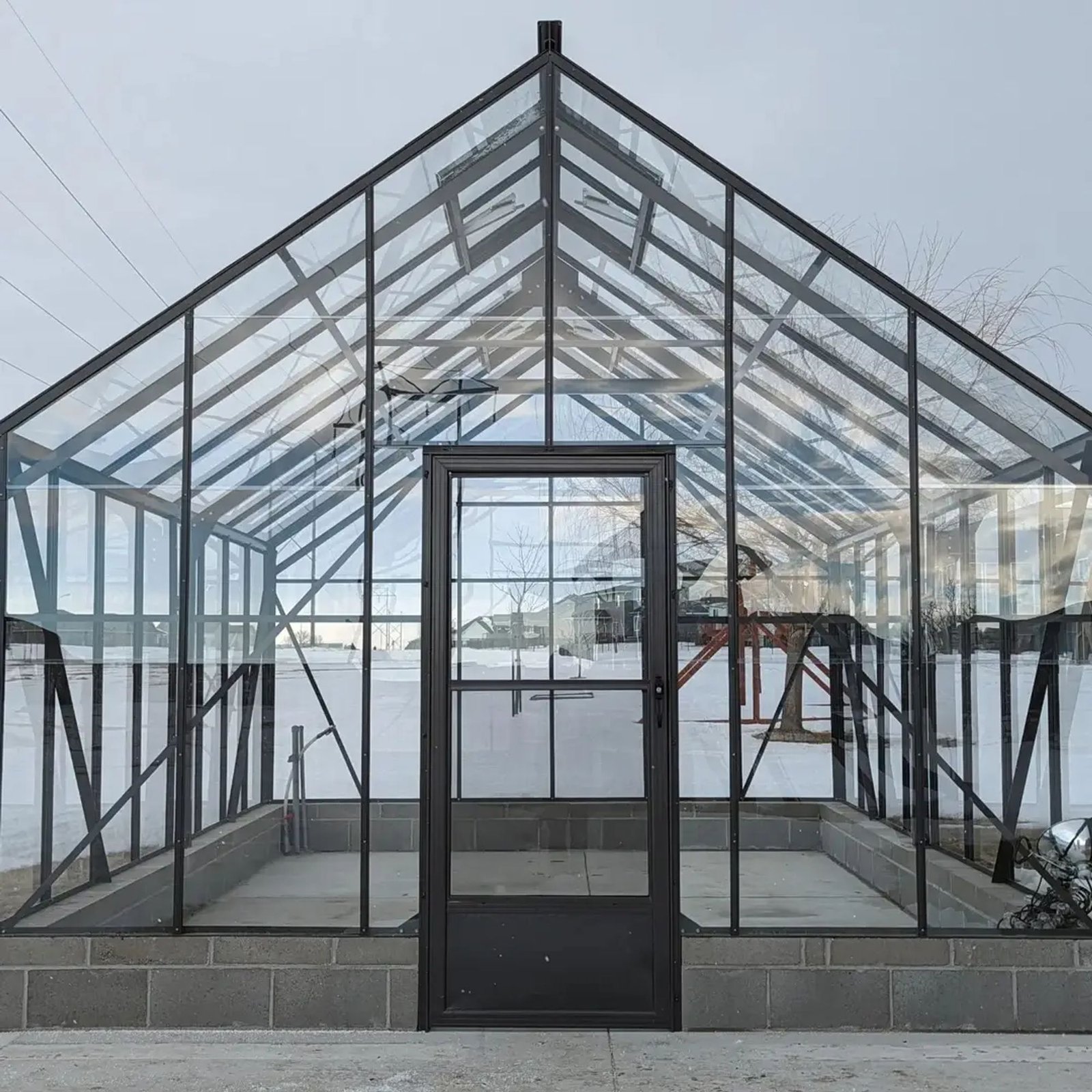Glass greenhouse with dark metal frame and central door, set on a concrete block base in a snowy yard