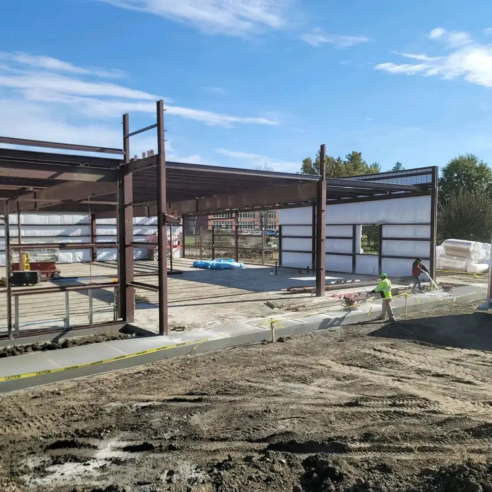 Two workers in high-visibility vests beside a steel-framed commercial building under construction, with fresh concrete walkway and dirt in foreground