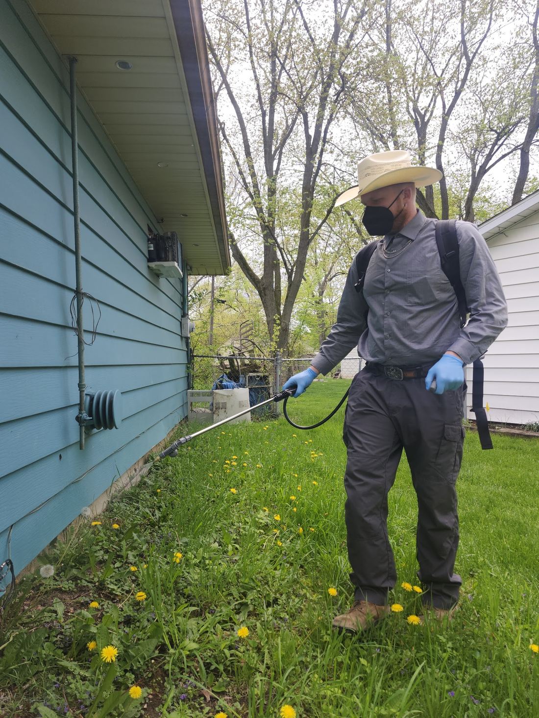 A man in a straw cowboy hat, black face mask and blue gloves sprays along the base of a blue house with a backpack sprayer