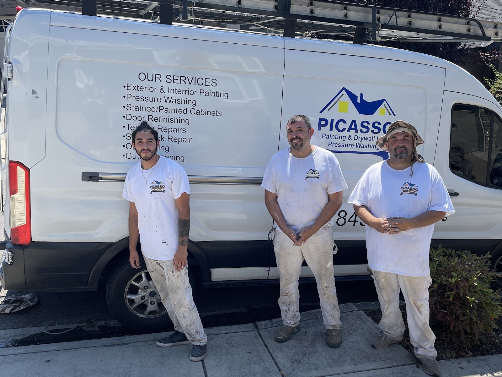 Three painters in paint-spattered white clothes stand in front of a white Picasso Painting & Drywall van with ladders on the roof