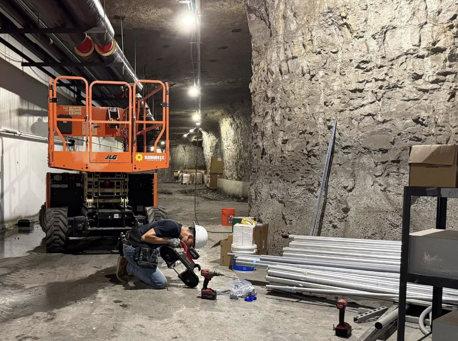 Electrician kneeling with a drill and cutting tool beside a scissor lift and stacked conduit in an underground space