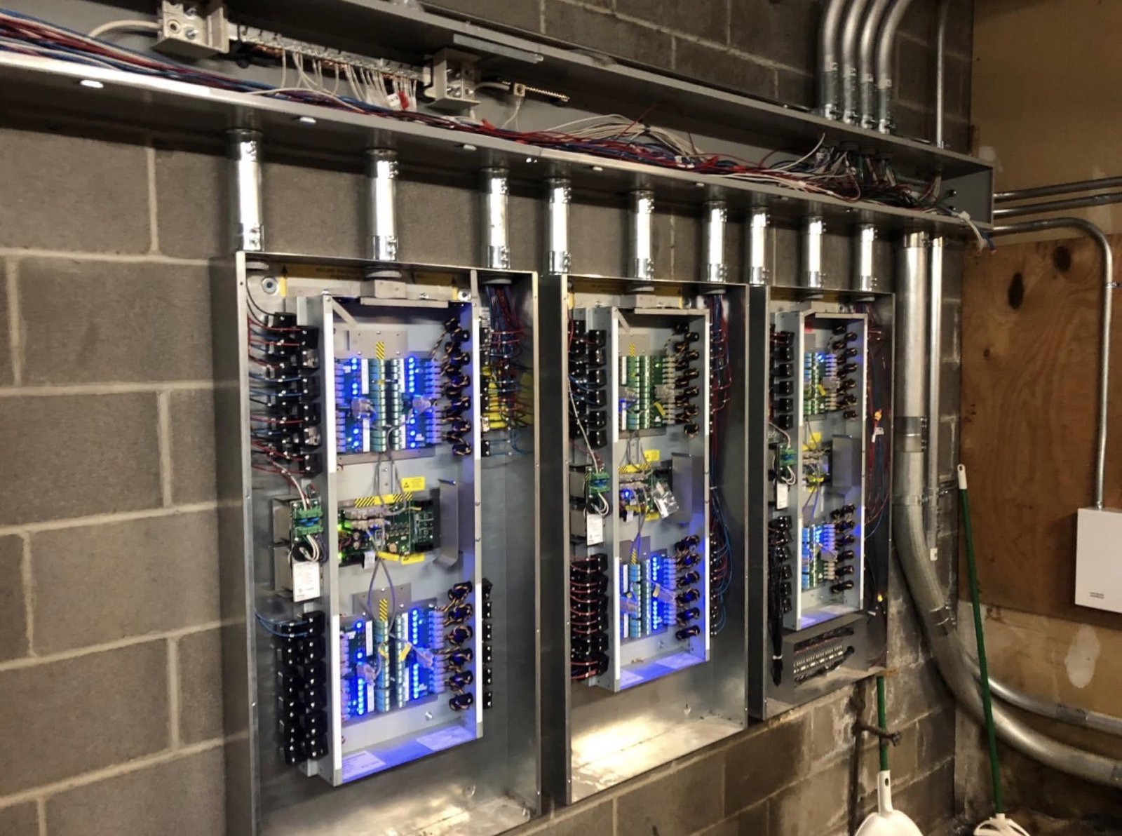 Three wall-mounted electrical control panels with blue-lit circuit boards and neatly routed conduit on a cinder block wall