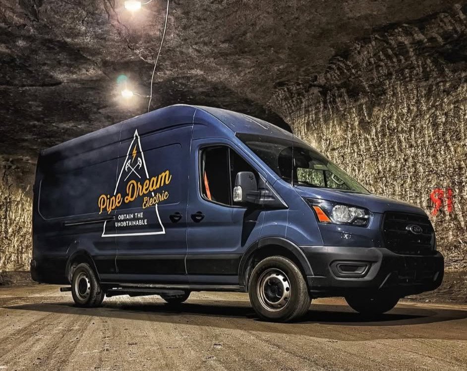 Navy Pipe Dream Electric work van with company logo parked inside a lit underground cave-like facility