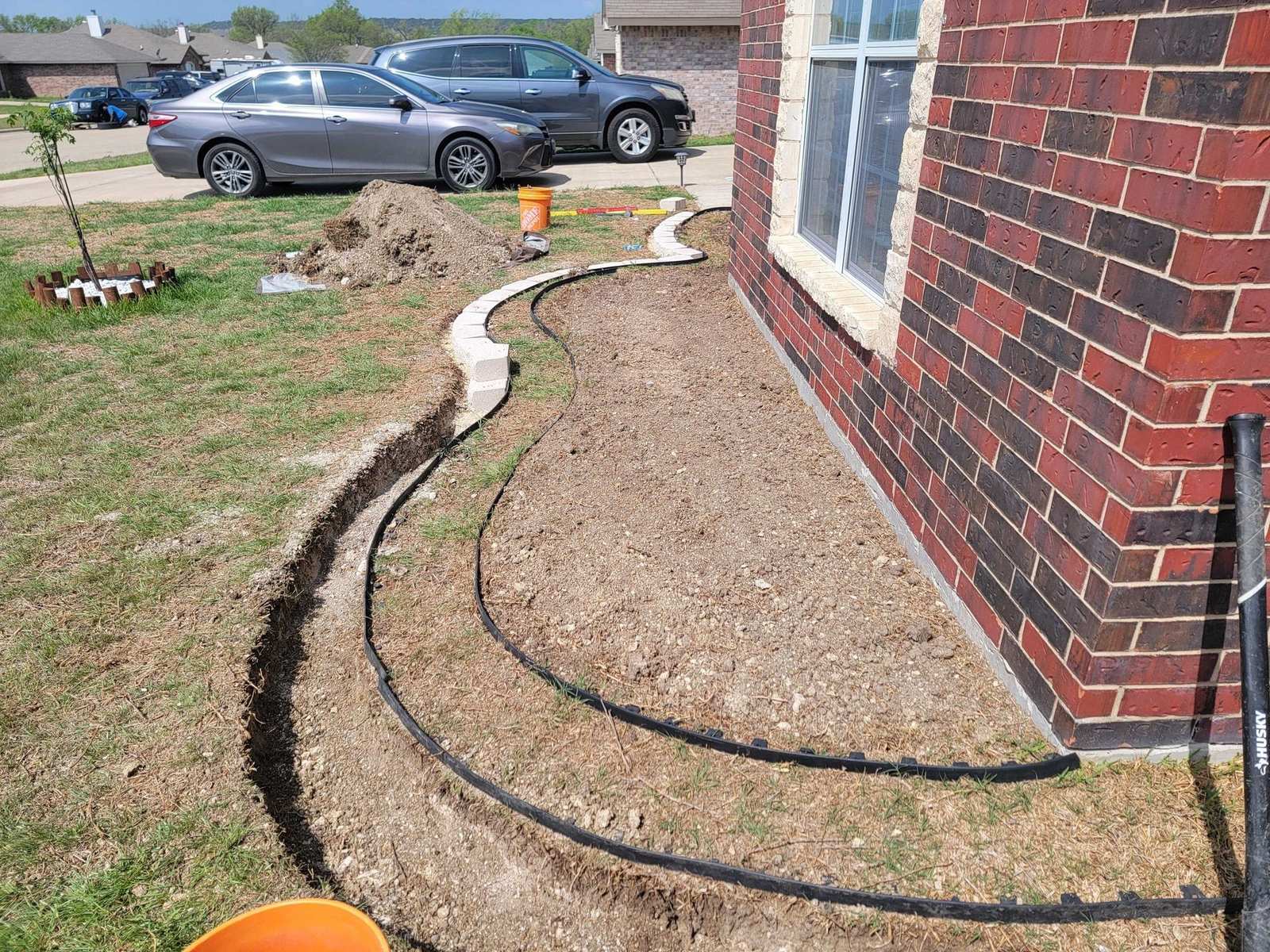 A curved bed border being installed in bare soil along a red brick house