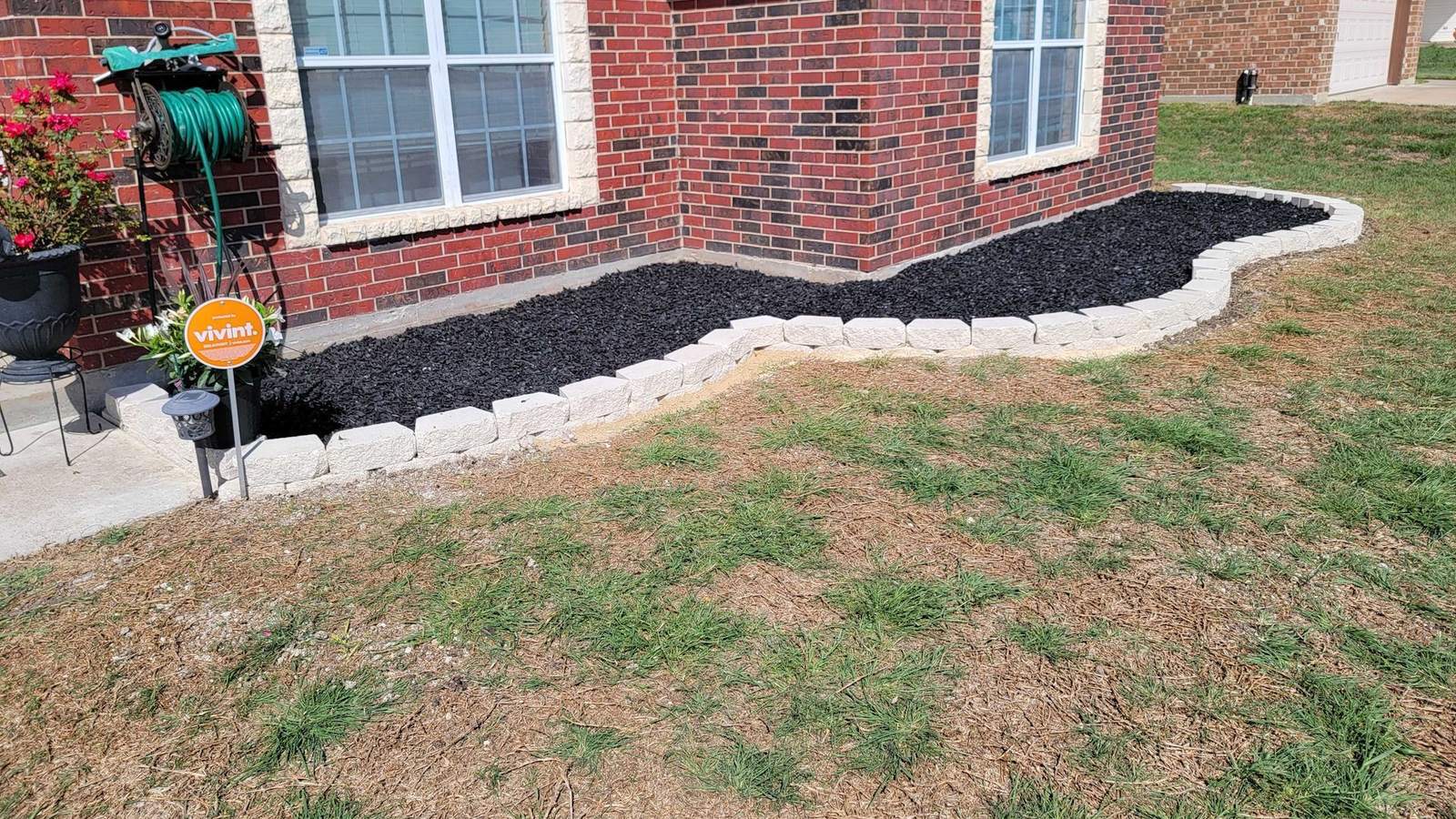 A finished flower bed with black mulch and a pale block border in front of a red brick house