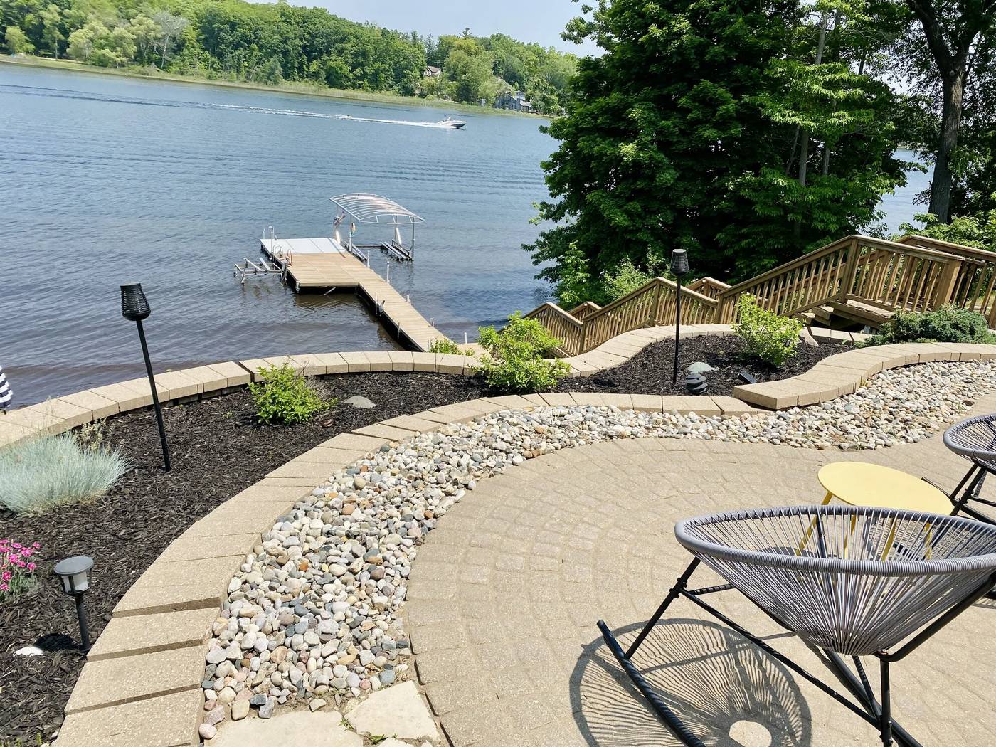 Mulch and river rock beds curving around a paver patio above a lake, with a dock and a boat on the water beyond