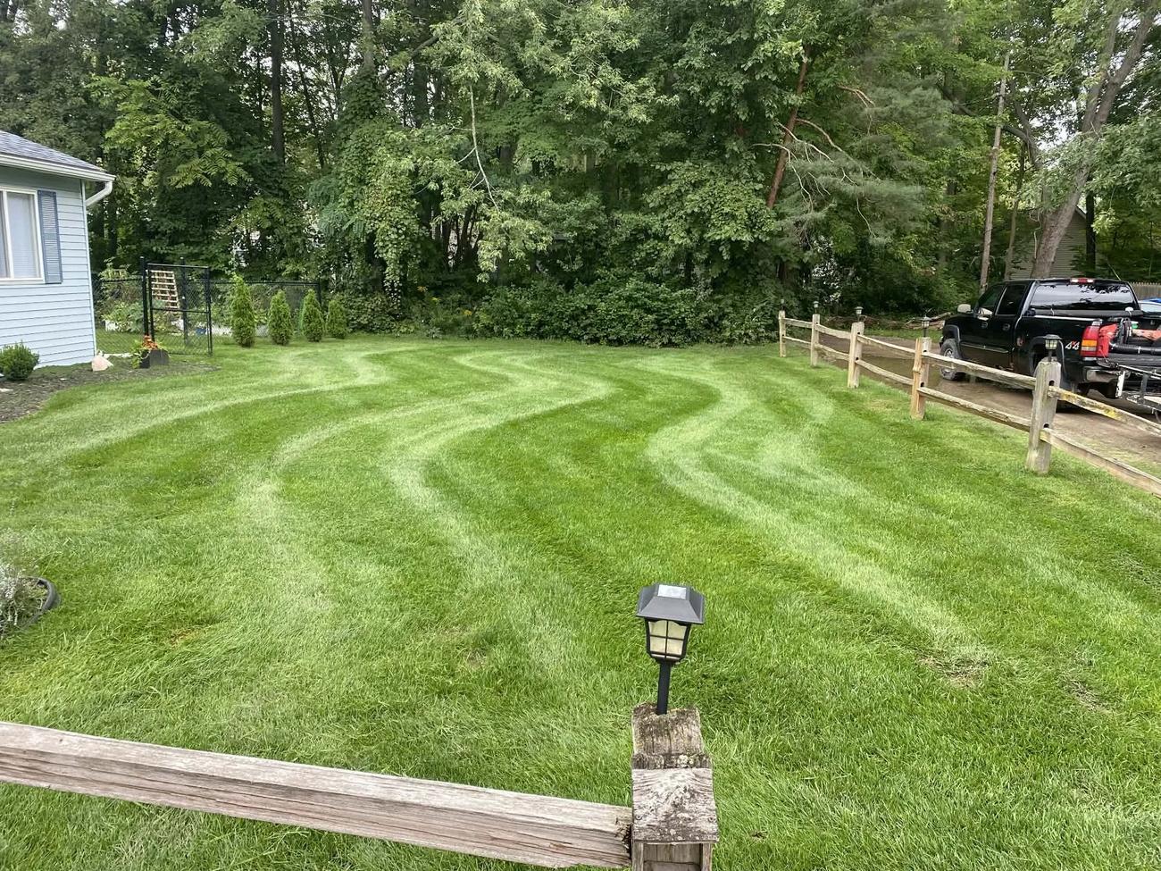 A lawn mowed in wavy stripes inside a split-rail fence, with a black pickup and trailer parked in the driveway beyond