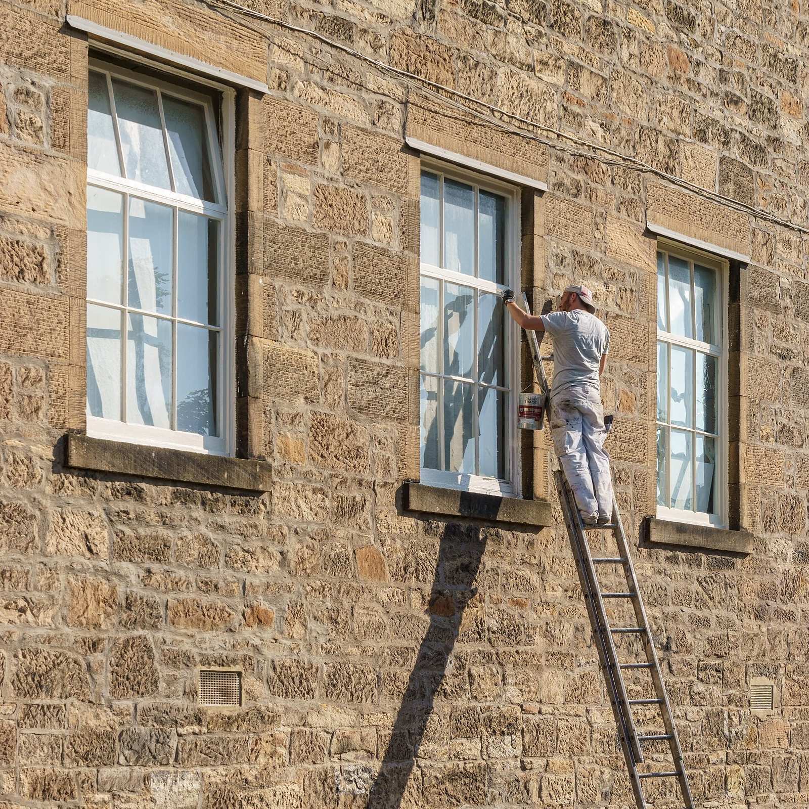 A painter on a tall ladder paints a white window frame on a sandstone building