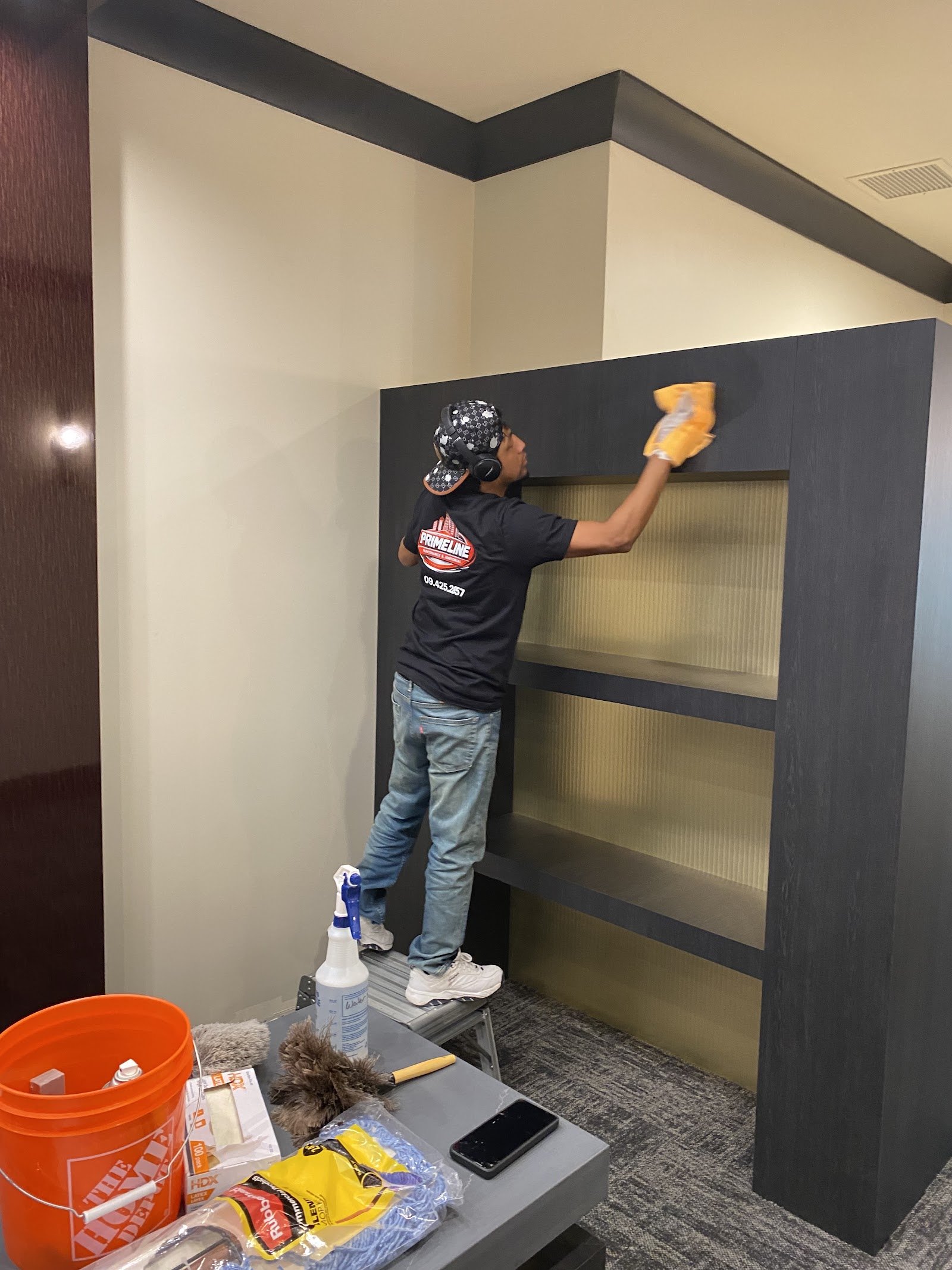 Worker in a Primeline shirt standing on a step stool wiping a dark built-in shelving unit in an office