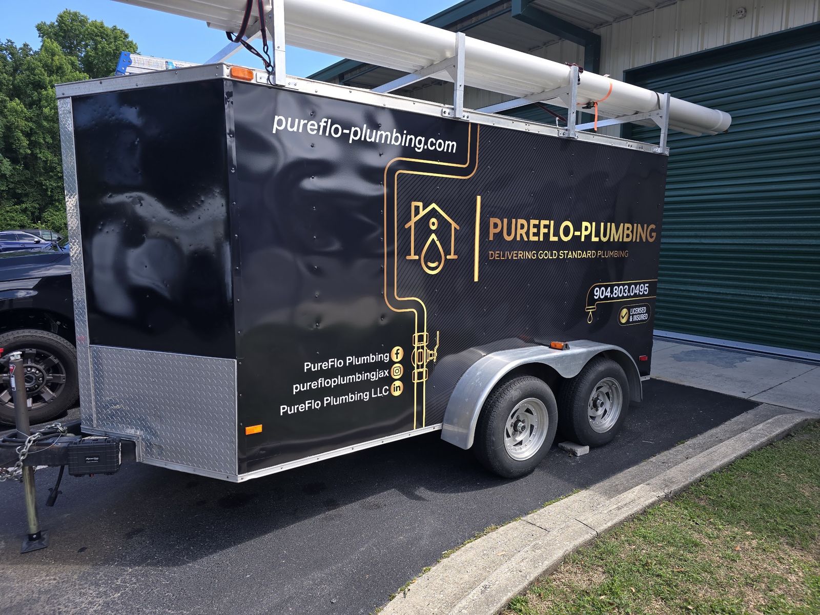 A black PureFlo Plumbing trailer with gold lettering and pipe racks, parked beside a metal building