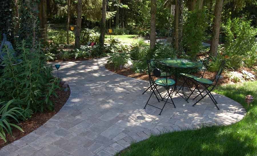 Curved gray paver patio with a green bistro table and chairs surrounded by mulched beds and trees