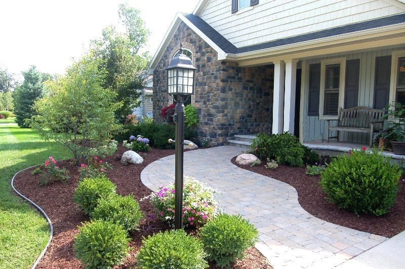 Curved paver walkway with a lamp post, mulched beds and shrubs leading to a stone-faced house entrance