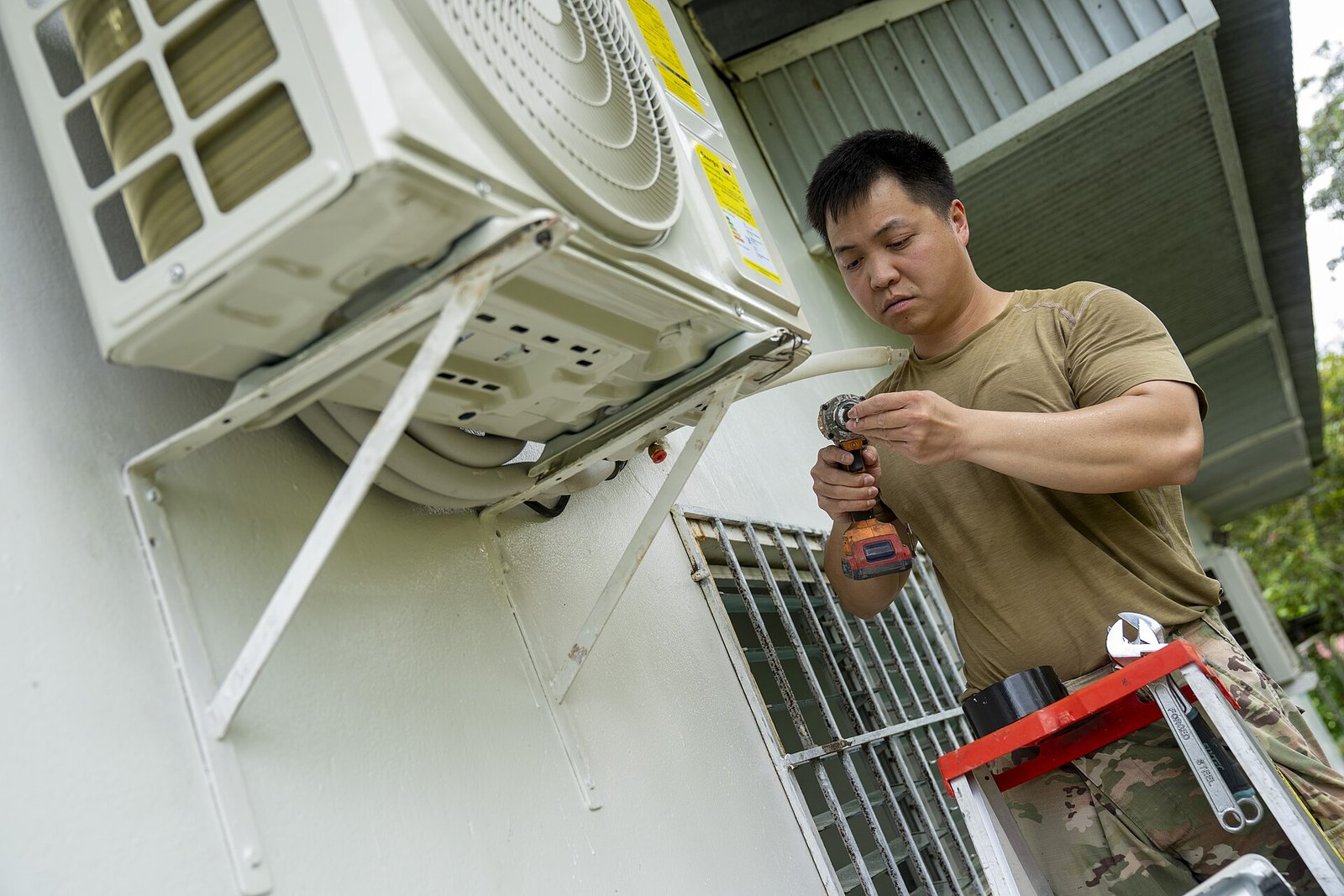 Technician in a tan shirt uses a cordless drill to secure a wall-mounted outdoor air conditioner unit on a bracket.