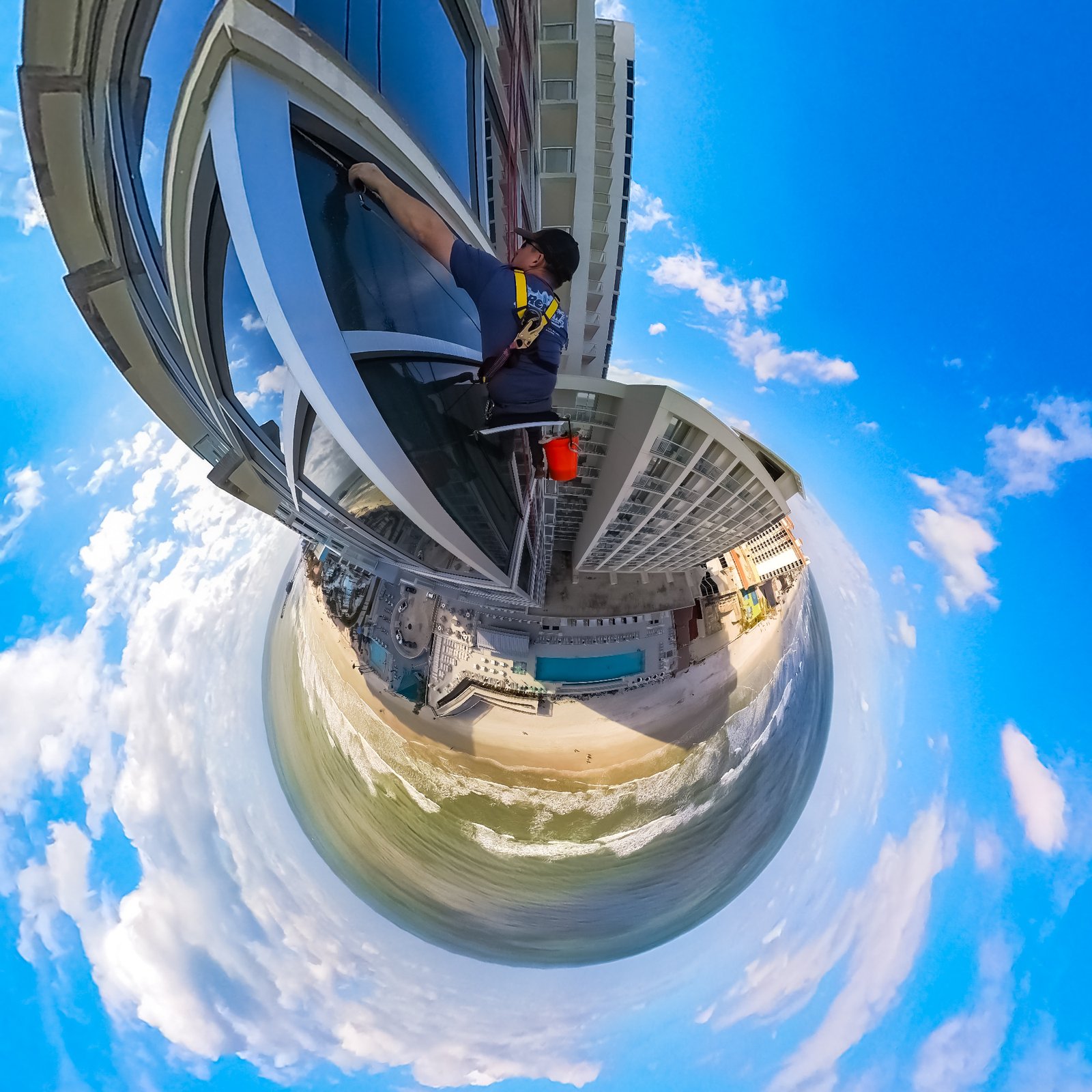 A window cleaner in a safety harness works on the glass of a tall oceanfront building, shown in a fisheye view.