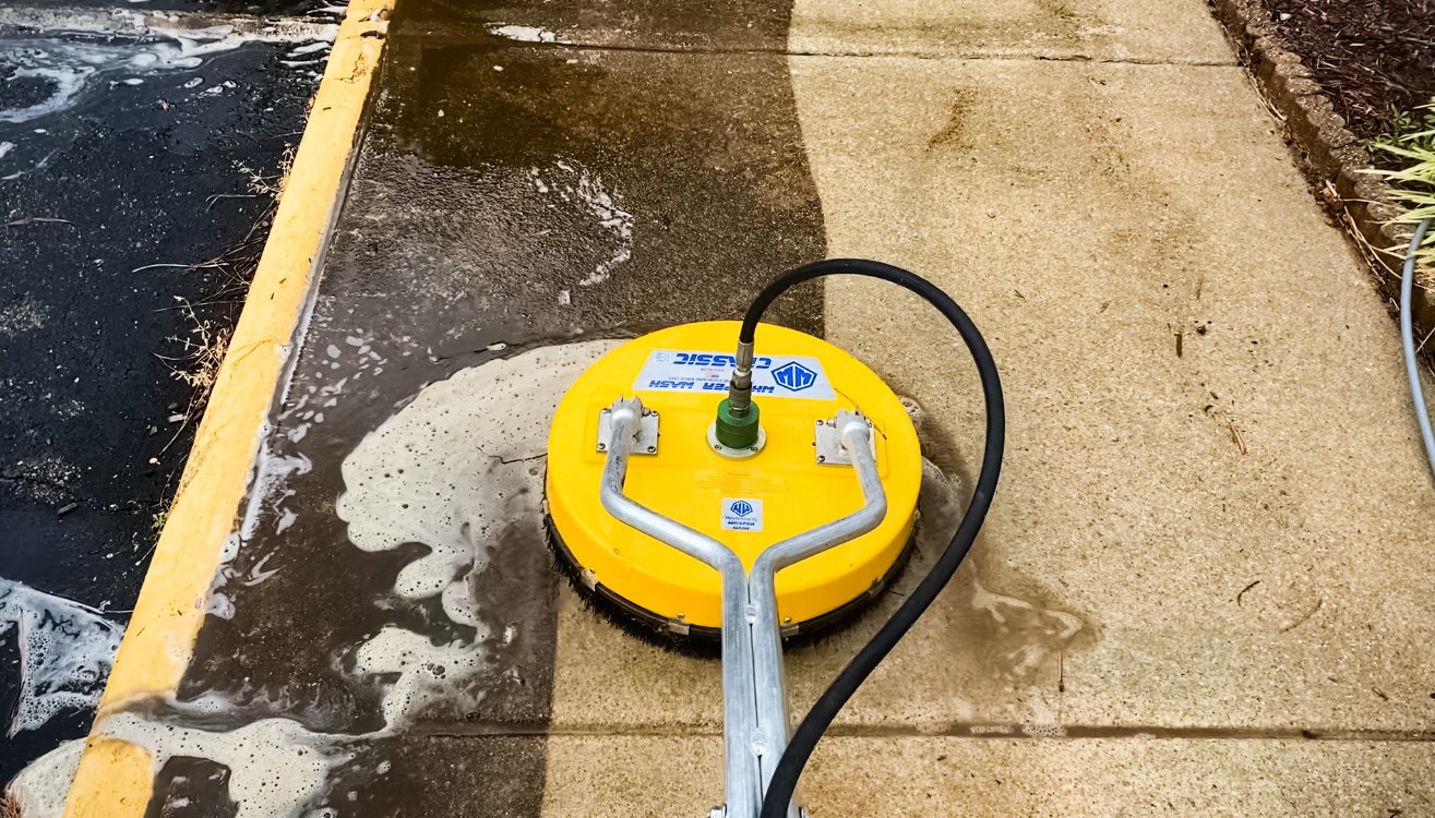 A yellow surface cleaner with a hose attached leaves a clean strip on a soapy, dirty concrete sidewalk beside a curb.