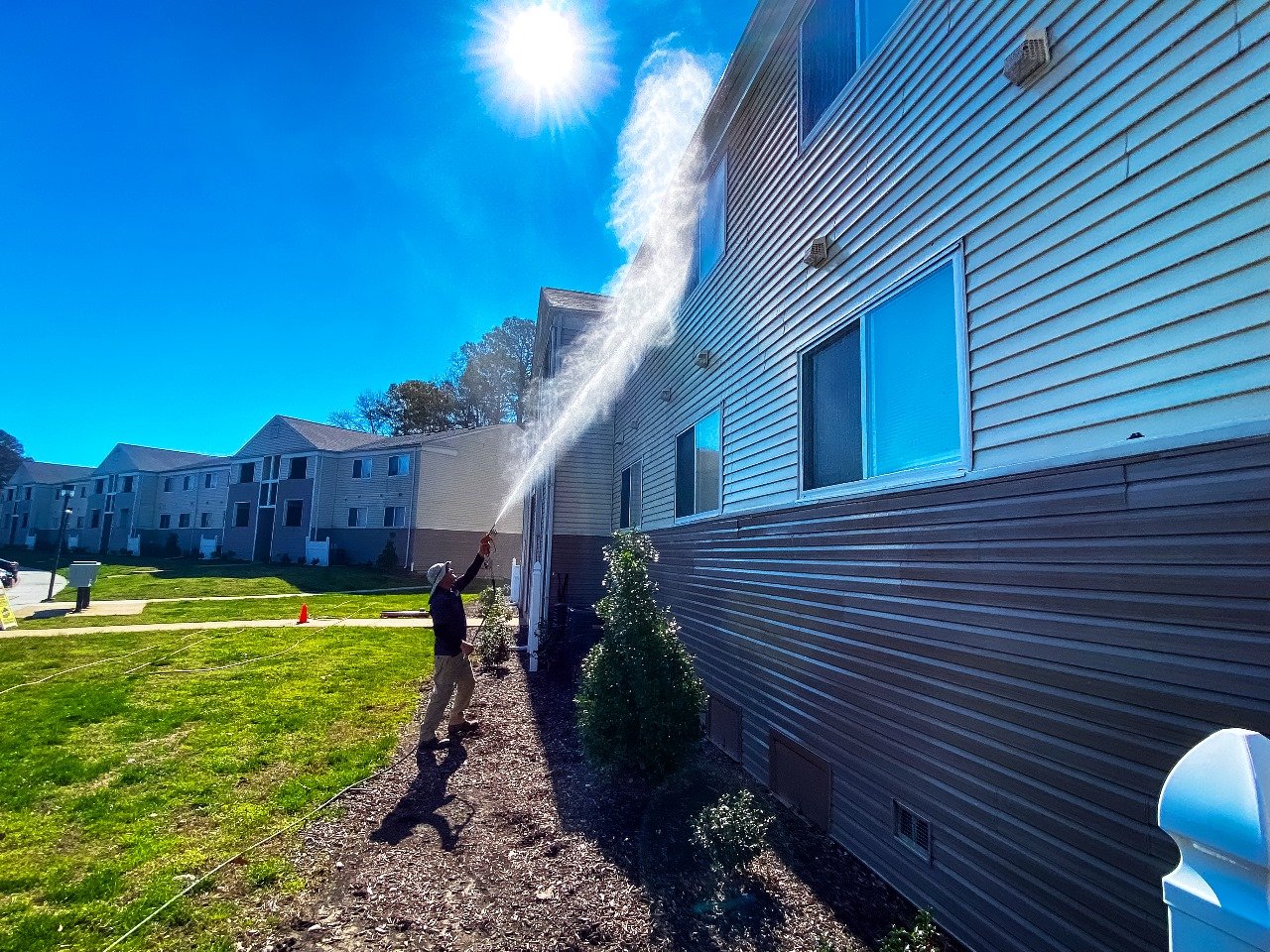 A worker sprays water up the vinyl siding of an apartment building on a sunny day, with more buildings beyond the lawn.