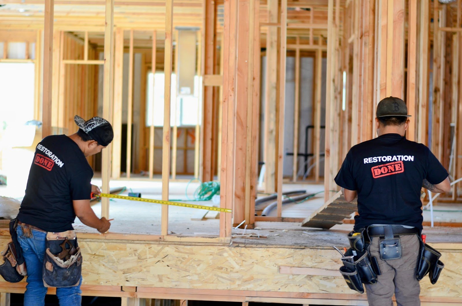 Two workers in black Restoration Done shirts and tool belts, one measuring with a tape, inside a wood-framed house