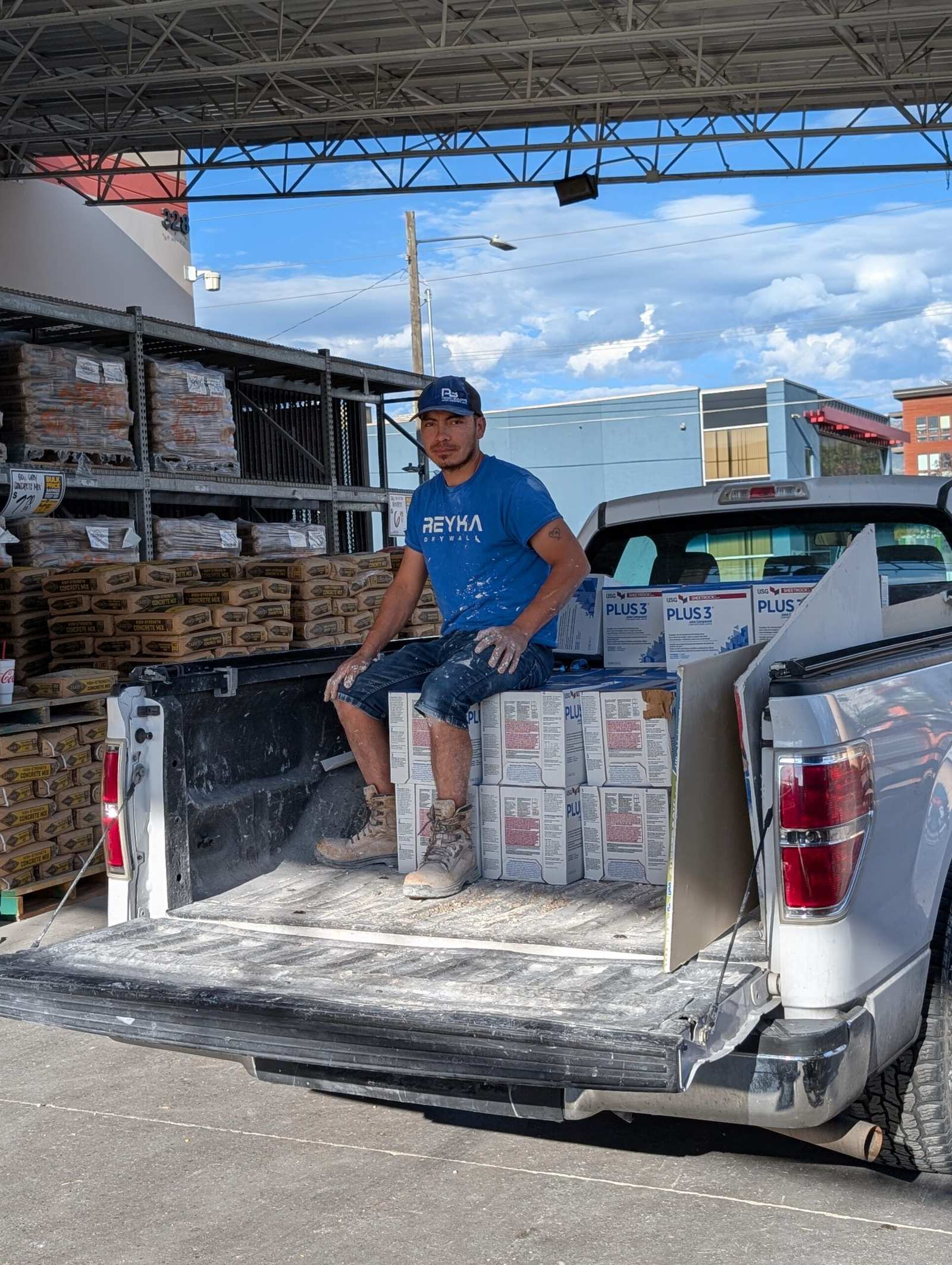 Worker in a Reyka shirt and cap sits on the tailgate of a pickup loaded with boxed drywall compound