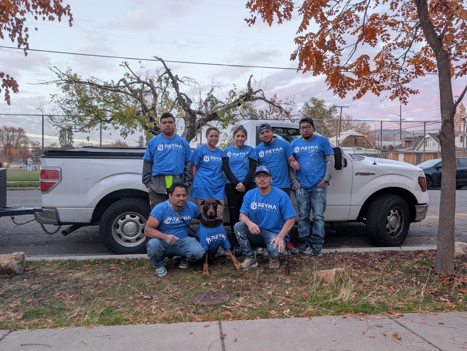 Reyka crew members in matching blue company shirts pose with a dog in front of a white pickup truck at dusk