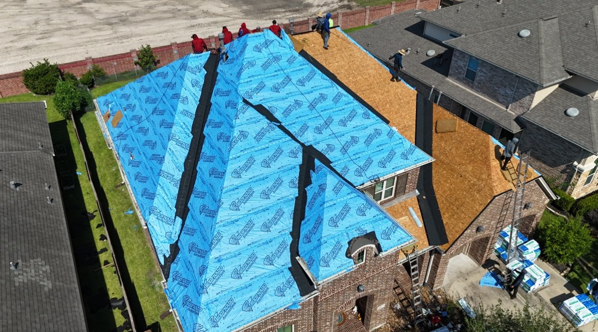 Aerial view of crew members working on a brick home's roof covered in blue underlayment, with shingle bundles staged nearby