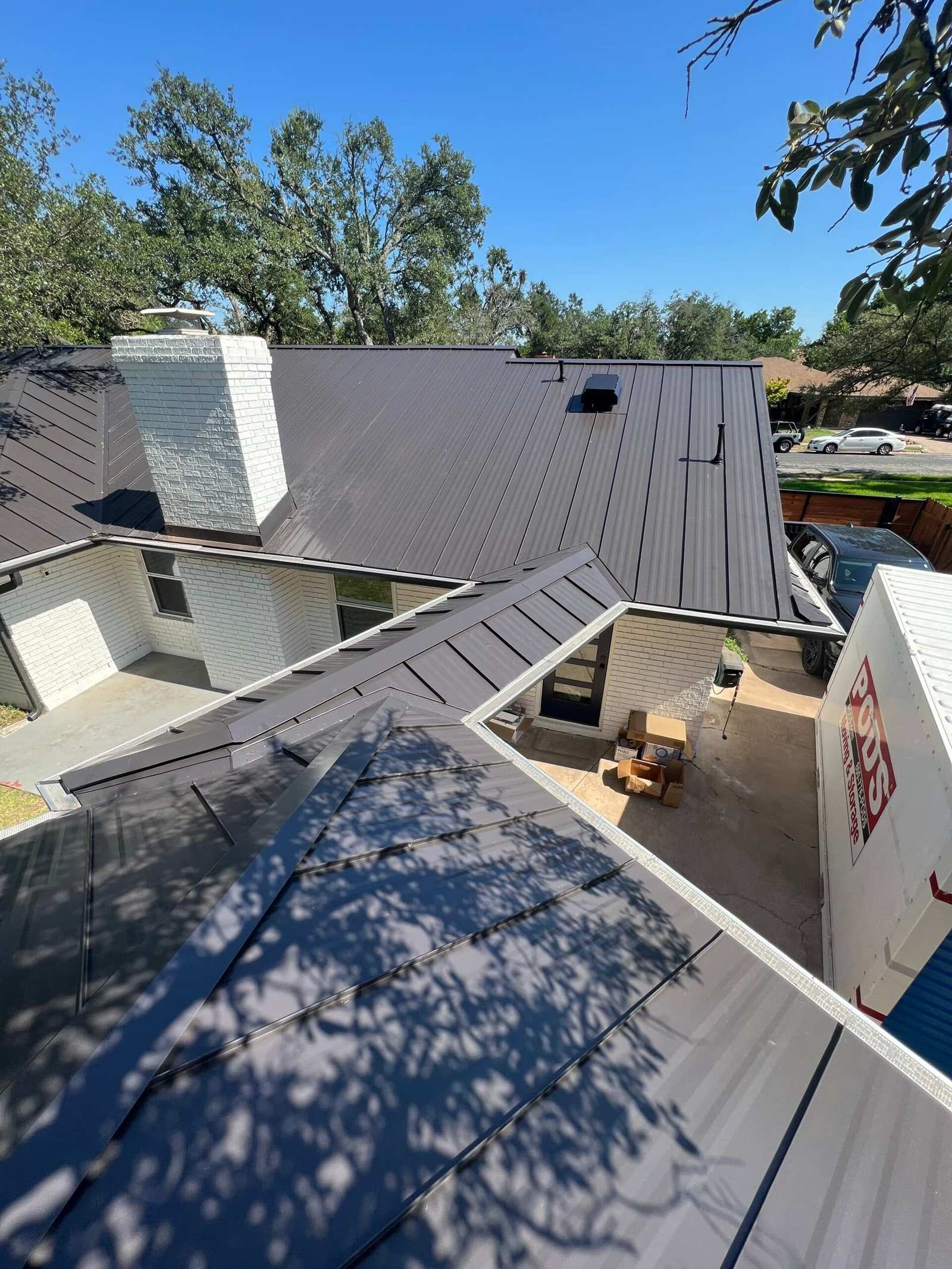Dark standing-seam metal roof on a white brick home with a chimney, seen from above with a container on the driveway