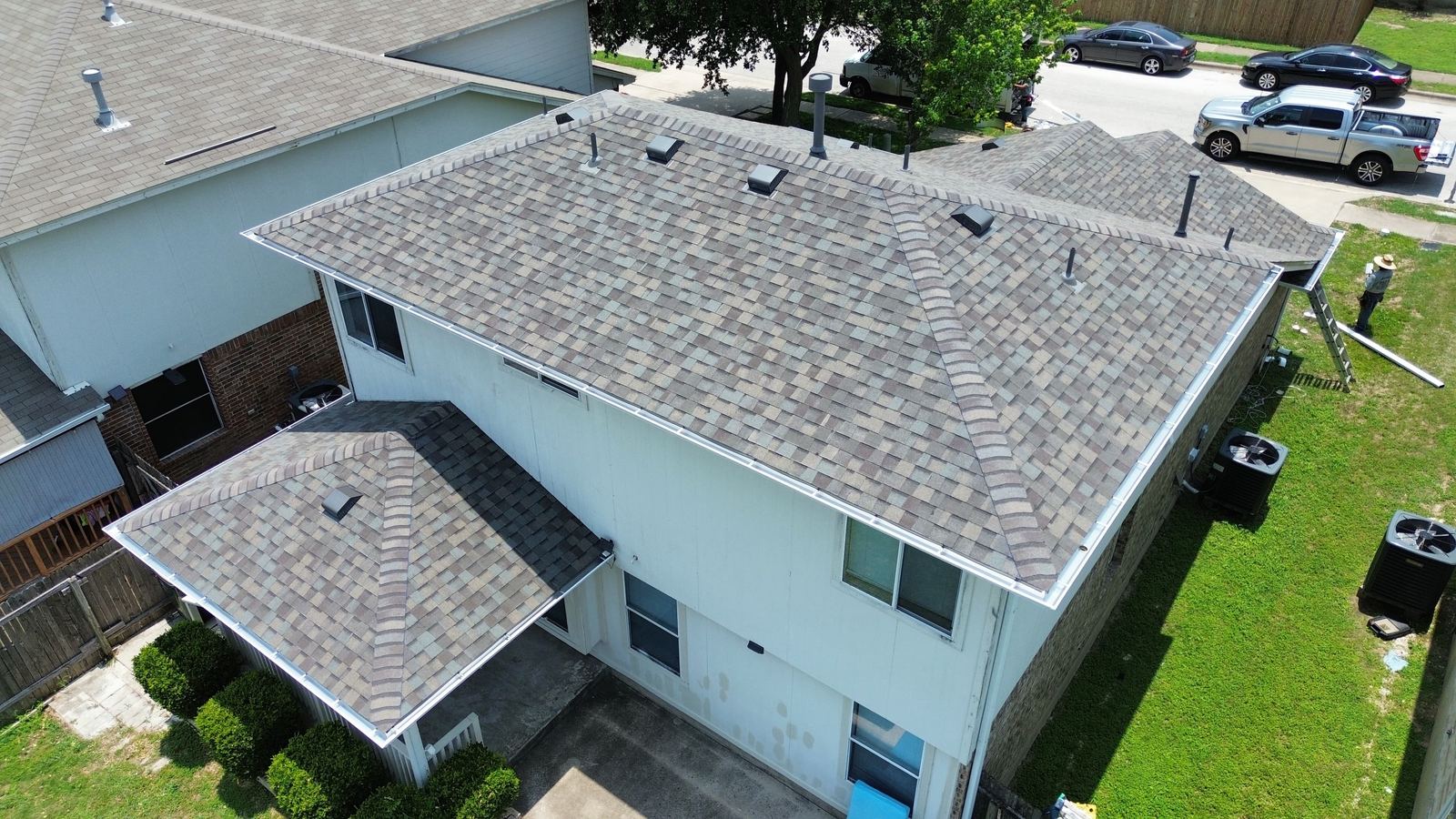 Aerial view of a two-story house with a new gray-brown shingle roof, vents and ridge caps, with trucks parked nearby