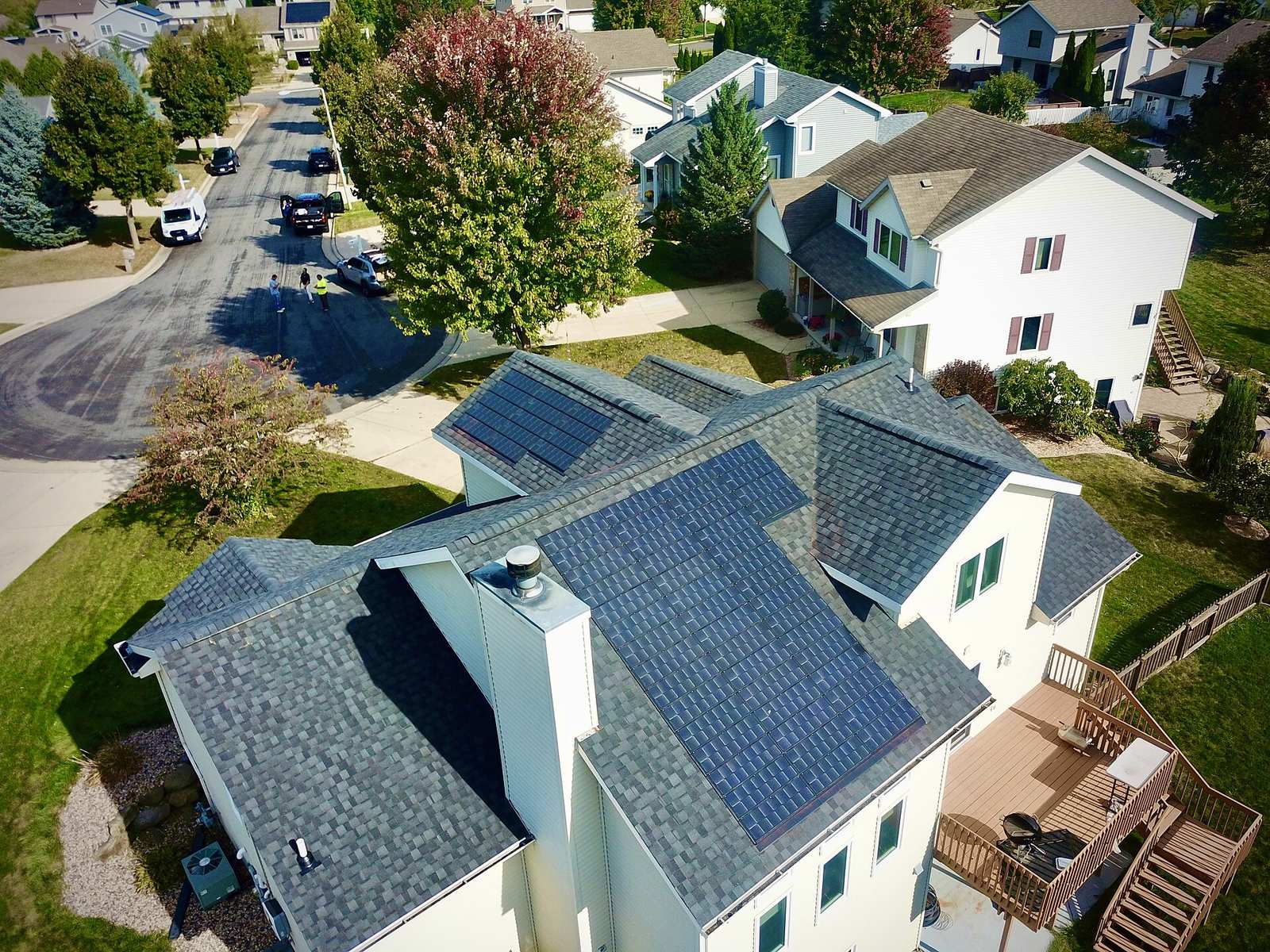 Aerial view of a suburban house with gray asphalt shingle roof, solar panels, chimney and wooden deck beside a residential street
