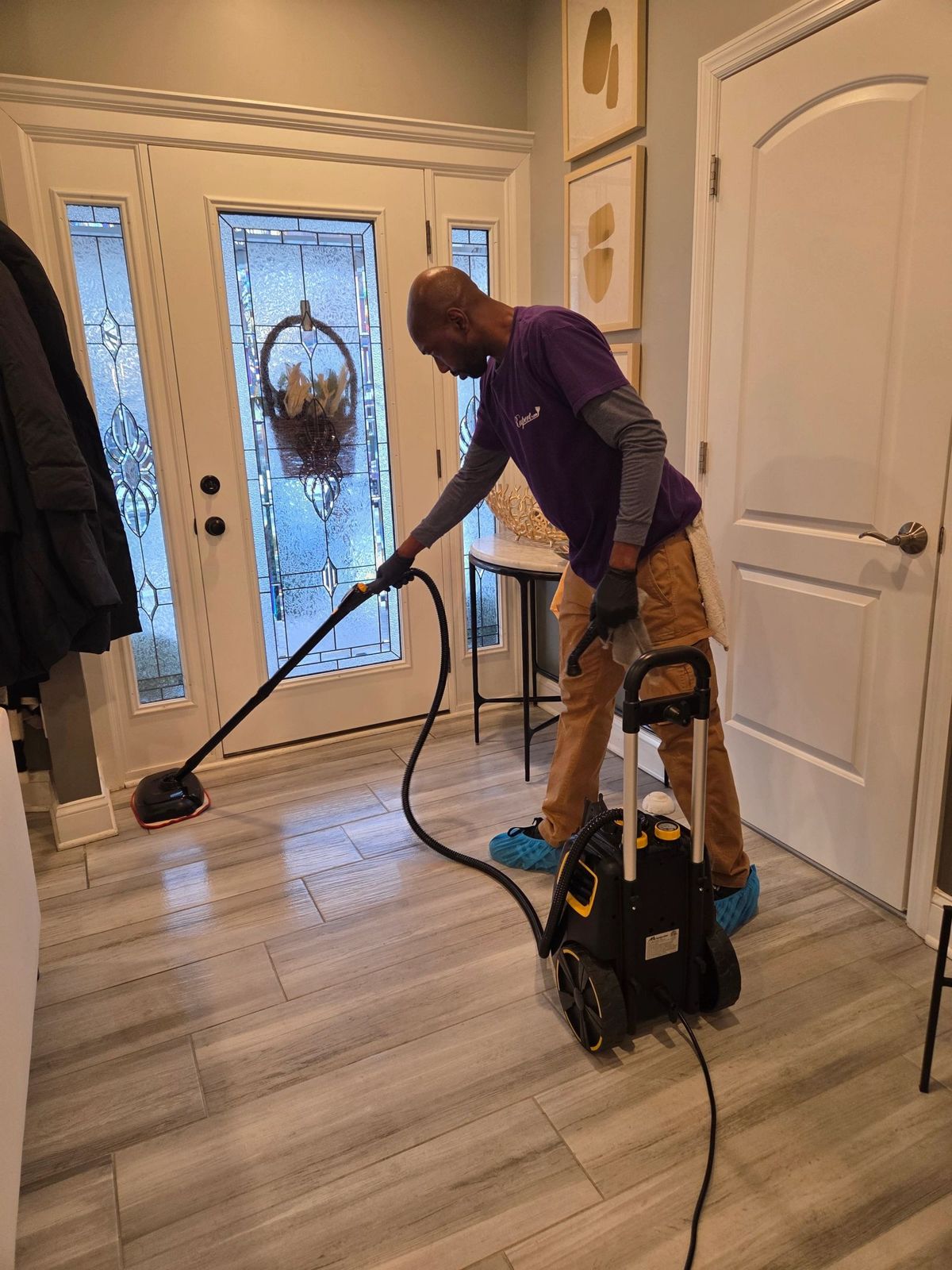 A cleaner in a purple shirt runs a canister steam cleaner across the tiled floor of a front entryway