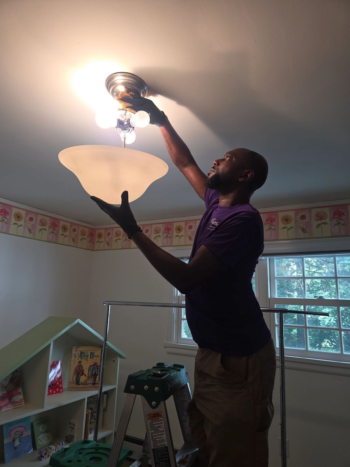 Rupert Richardson, in a purple shirt, cleans a ceiling light fixture in a bedroom, holding the glass shade in one hand