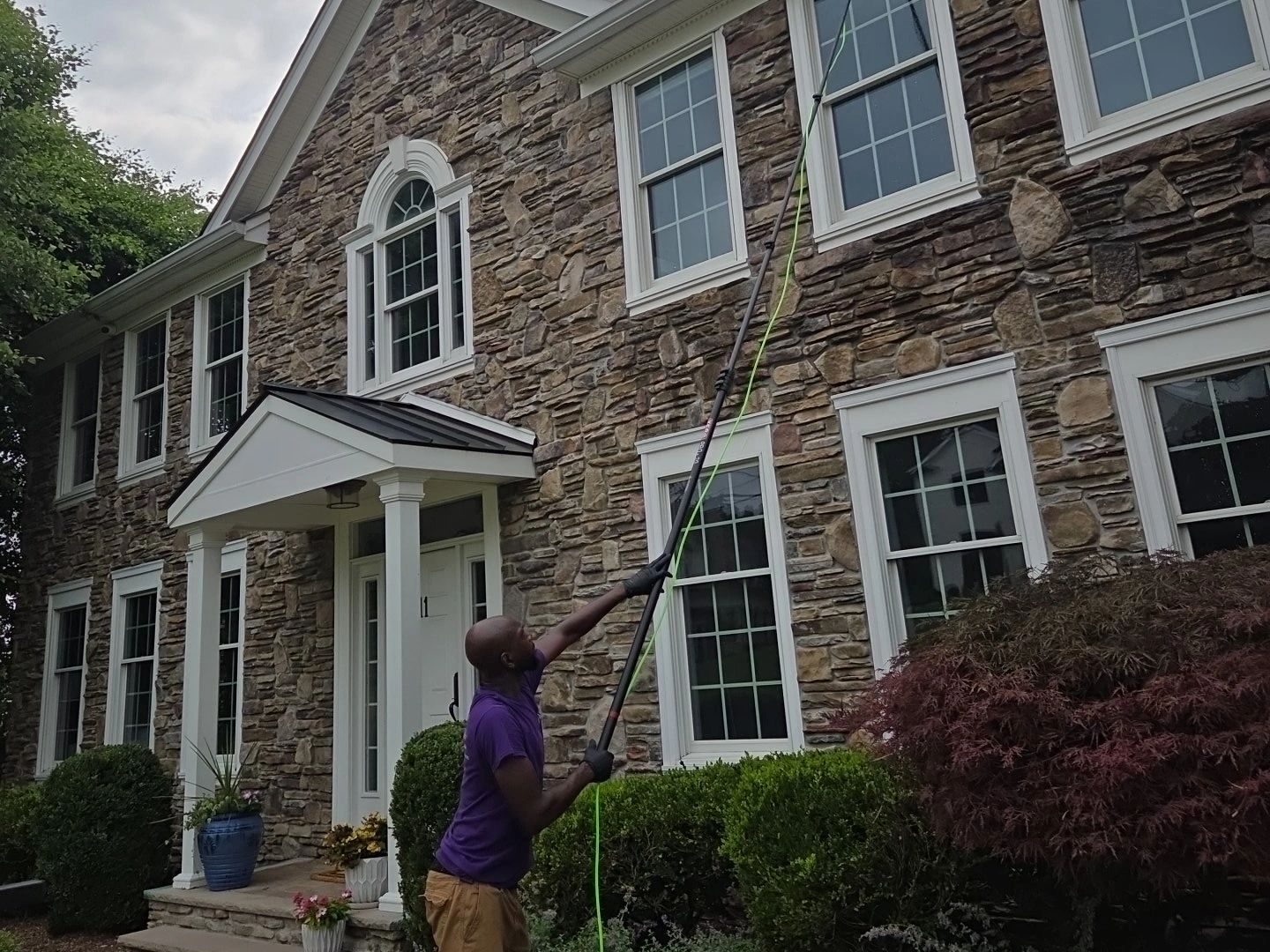 A cleaner in a purple shirt works a long water-fed pole against the upper windows of a two-story stone house