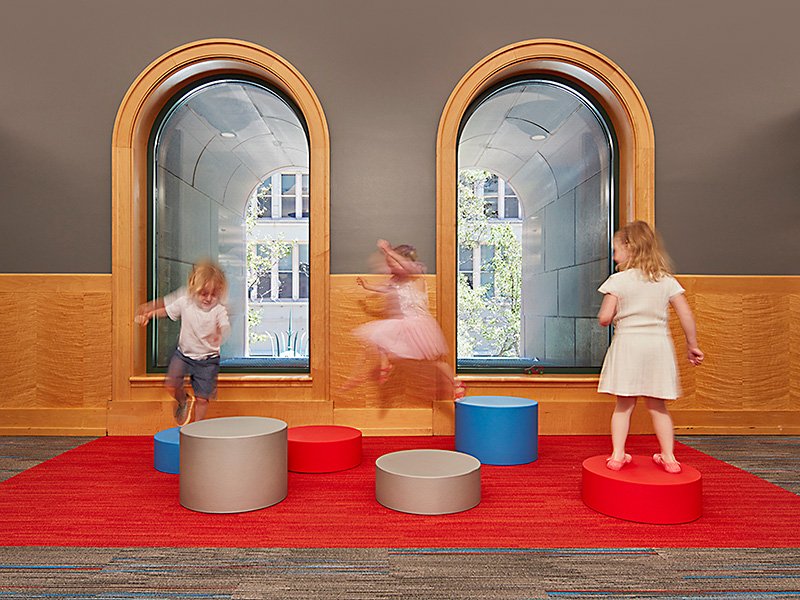 Children playing on round cushions atop a red and gray carpeted floor in a room with arched windows