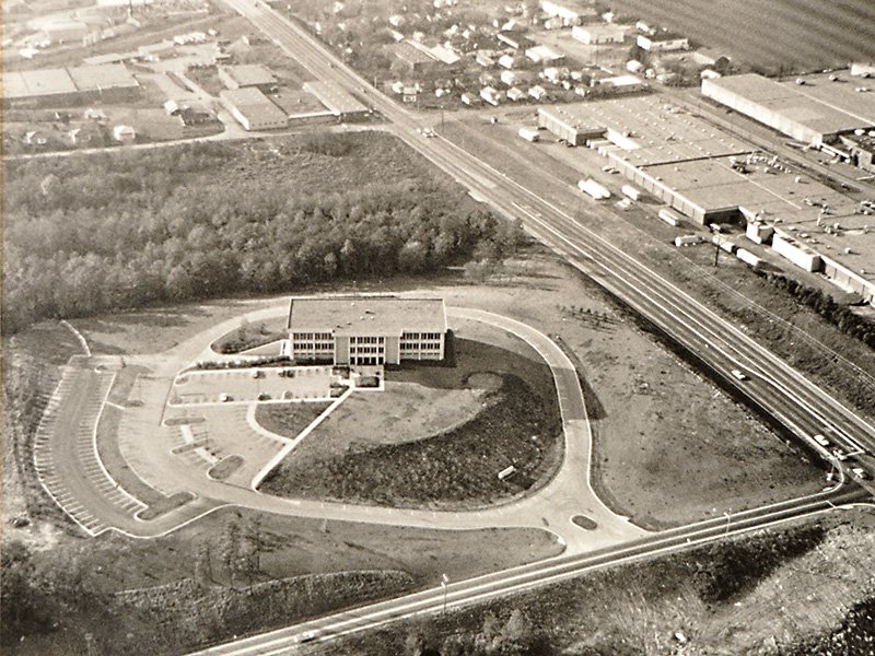 Black-and-white aerial view of a lone office building on a landscaped loop beside a highway and industrial buildings