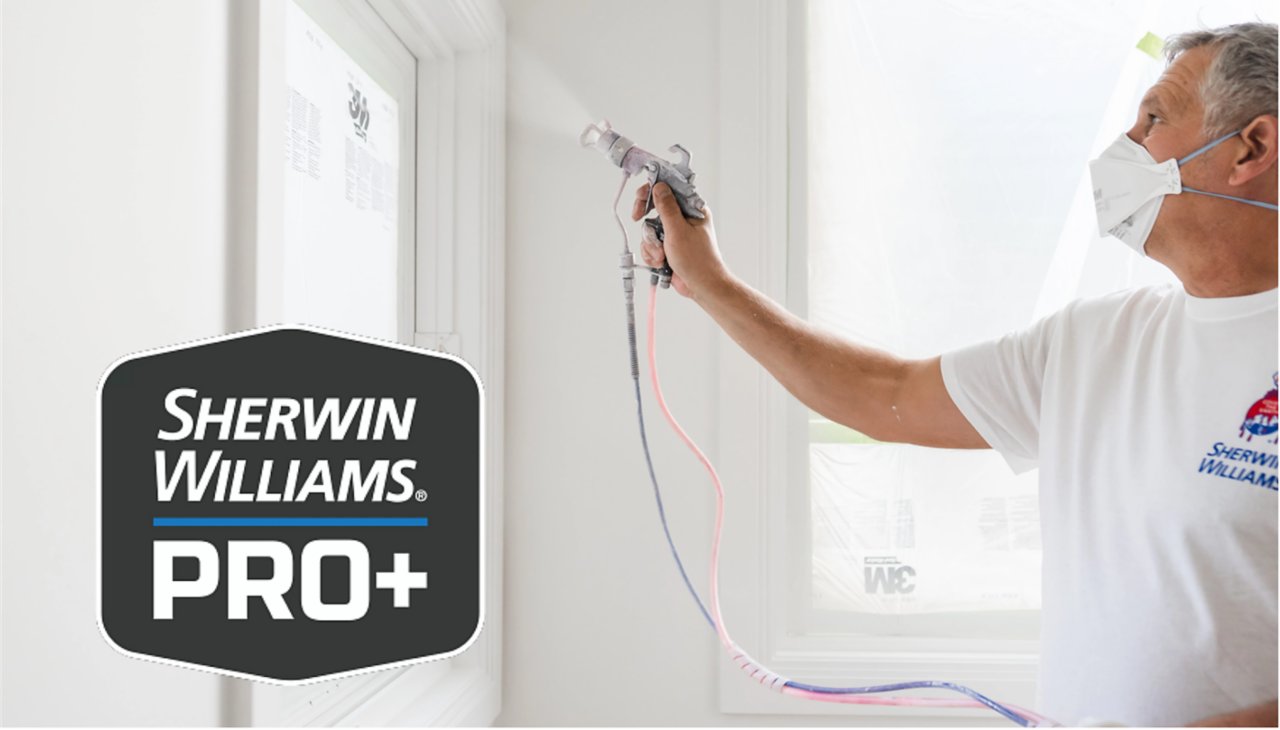 Painter in a mask and Sherwin-Williams shirt sprays paint on an interior wall beside a window covered in plastic