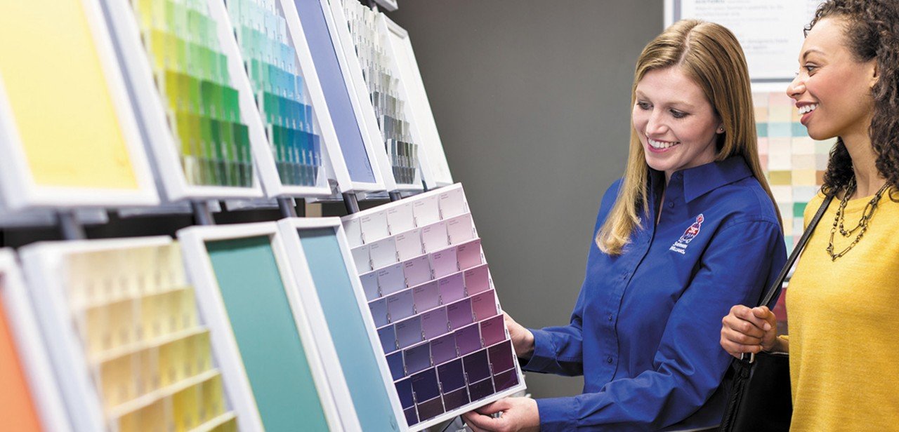 Sherwin-Williams employee in a blue branded shirt helps a smiling customer look over rows of paint color swatches in a store