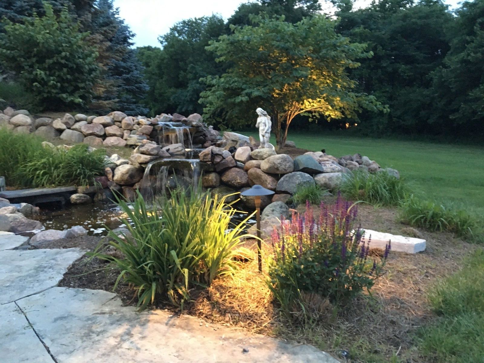 Boulder waterfall and small pond lit at dusk, with a statue, perennials and a flagstone walkway
