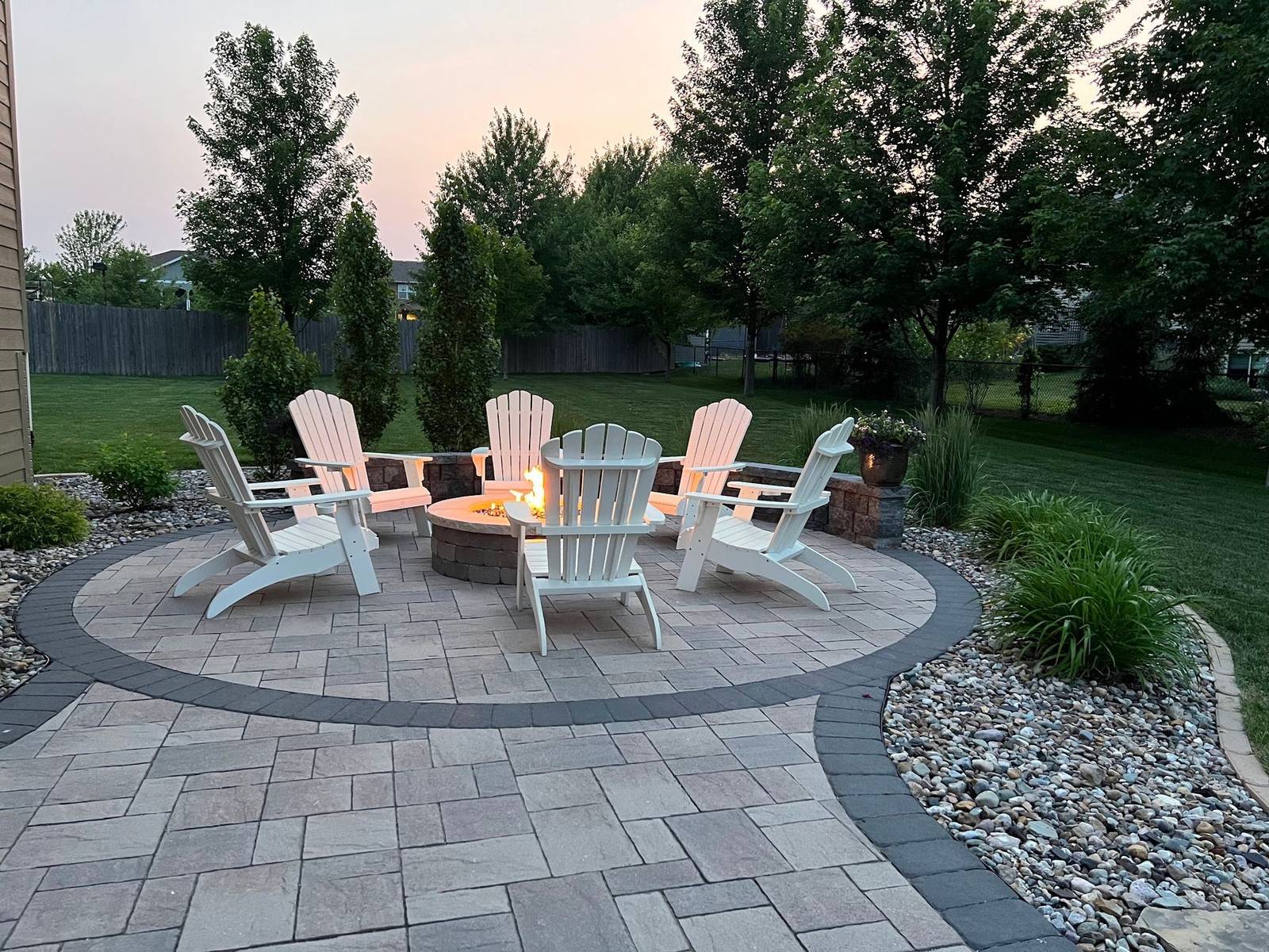 Paver patio with a round fire pit, six white Adirondack chairs and river rock beds at dusk